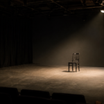 Empty black box theater stage with a single chair illuminated by a soft spotlight, representing space for reflection and possibility.